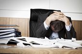 pic of working  - Worried businessman in dark suit sitting at office desk full with books and papers being overloaded with work - JPG 