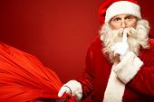 image of quiet  - Portrait of Santa Claus with huge red sack keeping forefinger by his mouth and looking at camera - JPG 