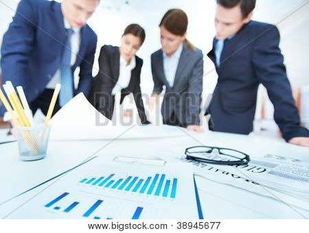 Picture or Photo of Business objects on background of engineers discussing blueprint at meeting